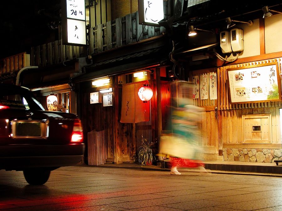 Una geisha cammina velocemente accanto a un taxi nel quartiere di Gion di notte.