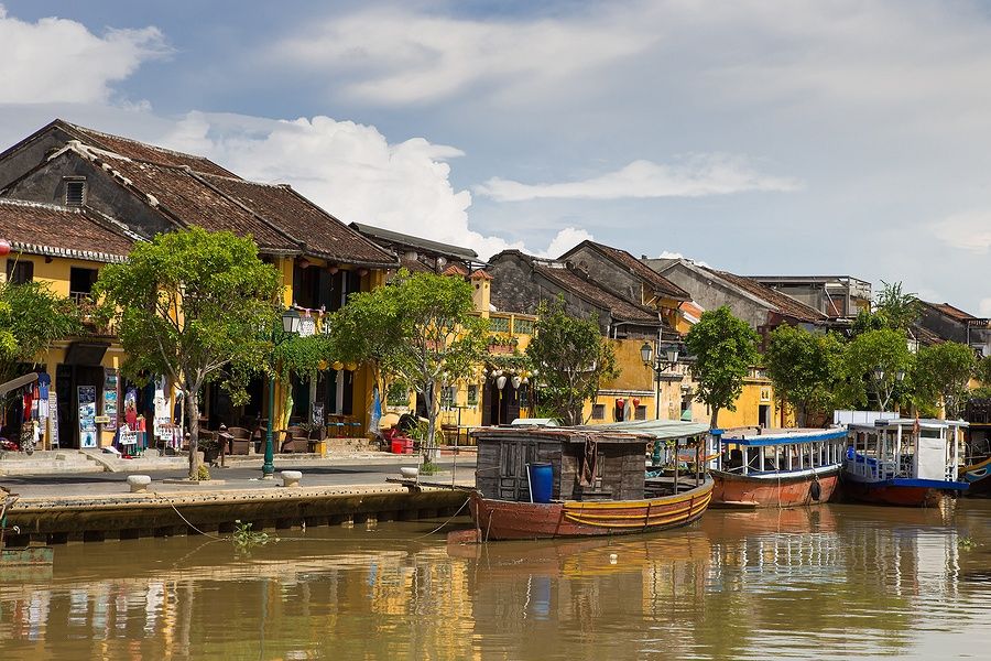 Barche di legno sul Fiume Thu Bon a Hoi An con case gialle storiche.