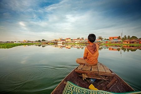 Ragazzo cambogiano sulla prua di una barca sul lago con villaggio nello sfondo.