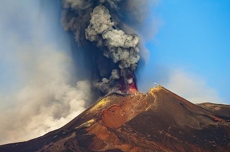 Eruzione del vulcano Etna con fumo e cenere nel cielo azzurro.