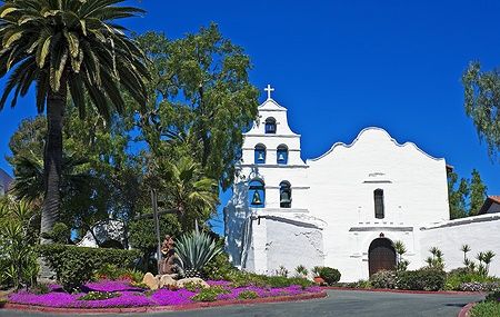 La Mission San Diego de Alcalá circondata da palme e fiori sotto un cielo blu intenso.