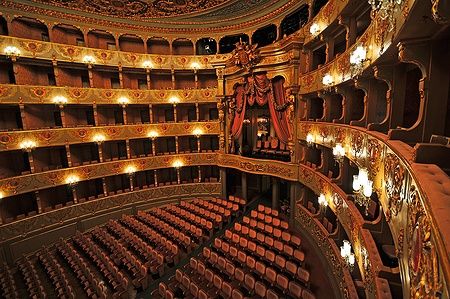 Interno di un teatro d'opera classico con balconate dorate e tende rosse.