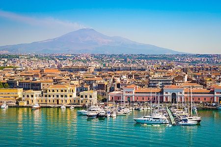 Vista del porto di Catania con barche, edifici storici e il vulcano Etna sullo sfondo.