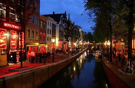 Strada illuminata ad Amsterdam di sera, con persone e canali.