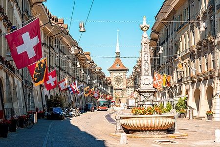 Veduta della Kramgasse a Berna con fontana e torre dell'orologio, bandiere svizzere lungo la via.