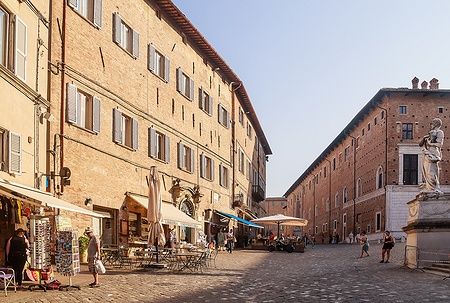 Piazza del Rinascimento a Urbino con edifici storici e persone che passeggiano.