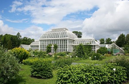 Serra del giardino botanico di Helsinki circondata da piante sotto un cielo parzialmente nuvoloso.