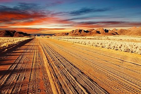 Strada polverosa nel Kalahari Desert, Namibia, con montagne al tramonto.
