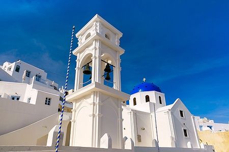 Chiesa Anastasi a Imerovigli, Santorini, con campanile bianco e cupola blu sotto un cielo limpido.