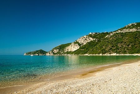 Spiaggia di ciottoli di Agios Giorgios Pagon con mare cristallino e colline verdi.