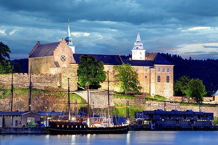 La Fortezza di Akershus illuminata di notte a Oslo con una barca a vela in primo piano.