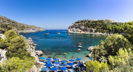 Baia di Anthony Quinn a Rodi con mare cristallino e ombrelloni blu.