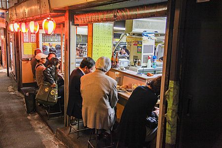Persone sedute al bancone di un piccolo locale lungo una strada stretta a Tokyo con lanterne rosse.