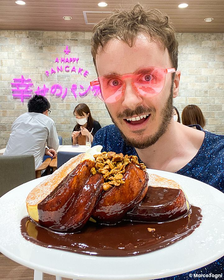 Marco Togni con occhiali rosa sorride tenendo un piatto di a Happy Pancake al cioccolato in un ristorante moderno.