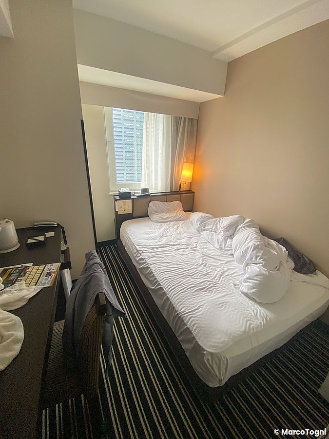 Interno di una camera compatta presso l'Apa Hotel a Shinjuku con letto singolo disfatto e finestra urbana.