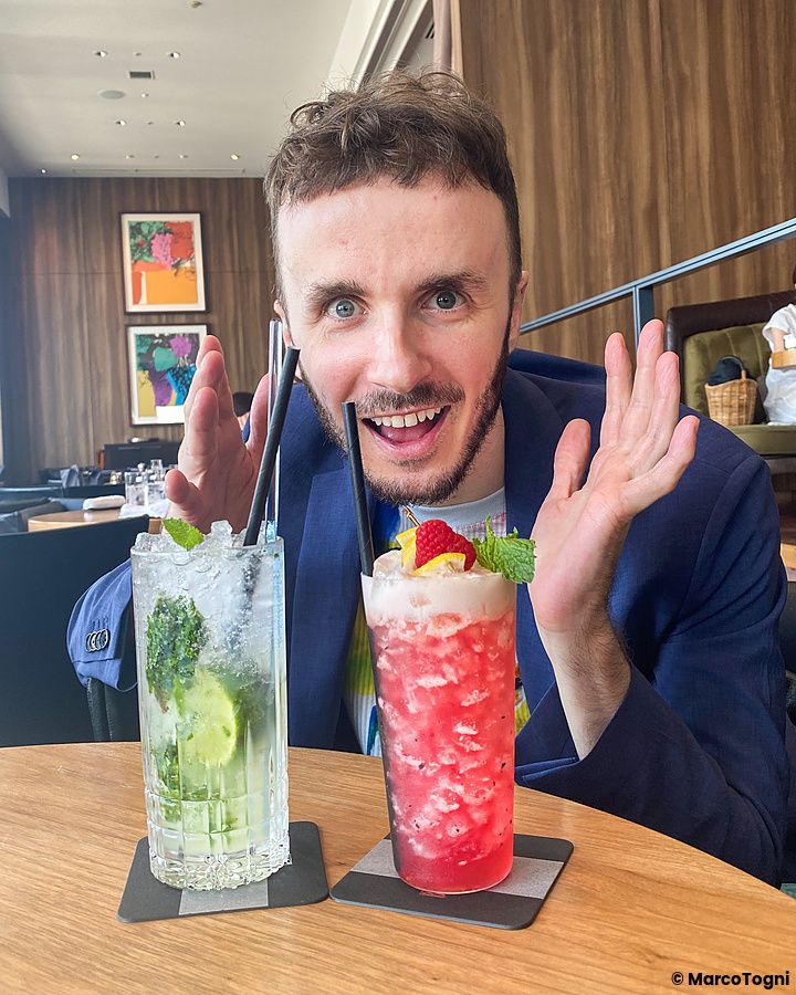 Marco Togni sorridente posa al bar Bellovisto a Shibuya, Tokyo, con due cocktail colorati davanti a sé su un tavolo.