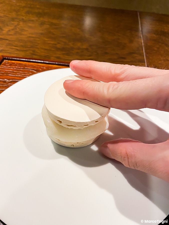 Un daifuku freddo con crema inserito tra due meringhe, tenuto fermo da una mano, servito su un piatto presso Denkushiflori.