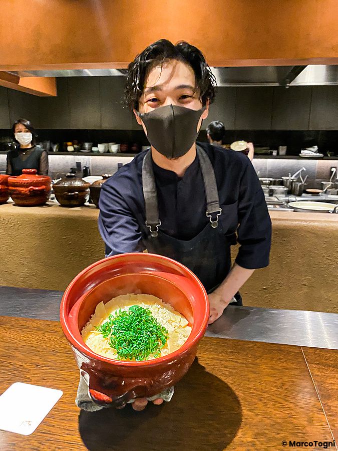 Uno chef indossa una mascherina e presenta un recipiente in ceramica contenente riso con takenoko al Denkushiflori.