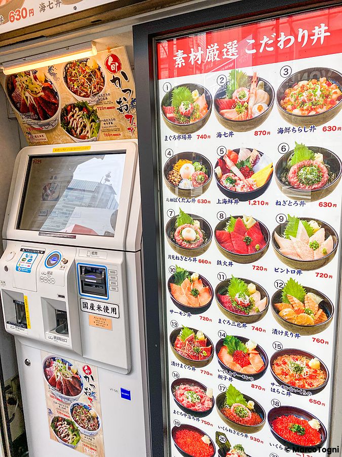Un totem automatico per l'ordinazione di cibi affiancato da un menu fotografico con venti ciotole di pesce e riso.