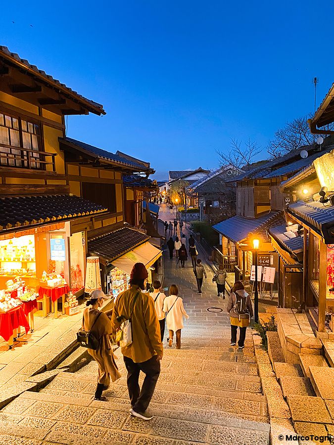 Persone camminano lungo la scalinata in pietra di Sannenzaka a Kyoto circondate da edifici tradizionali illuminati.