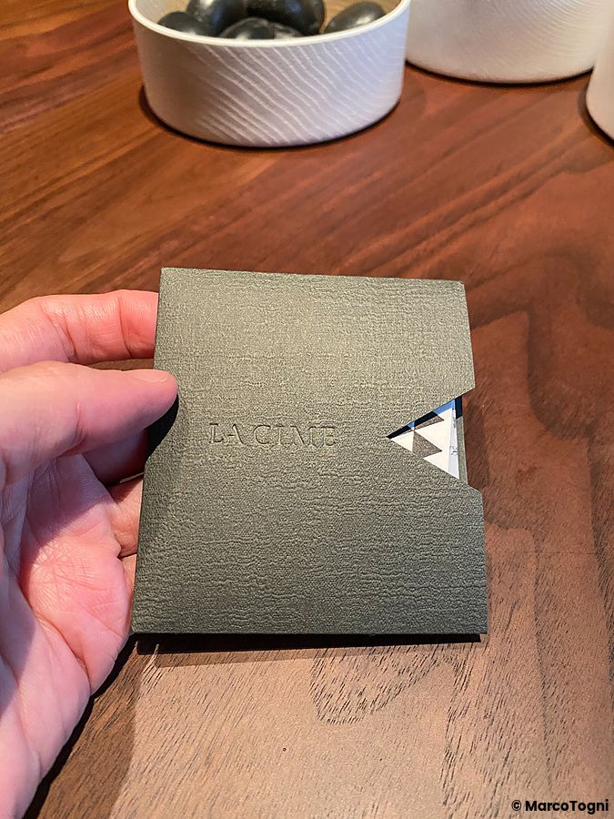 Una mano sostiene un porta menu grigio scuro con il logo del ristorante La Cime su un tavolo di legno.