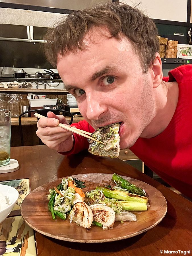 Marco Togni con un maglione rosso mangia del pesce con le bacchette seduto al tavolo del Ristorante Momose Shokudo.