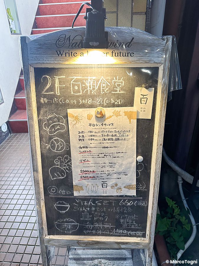 Lavagna pubblicitaria del Ristorante Momose Shokudo posta all'esterno, con menu scritto a mano e indicazioni sugli orari.