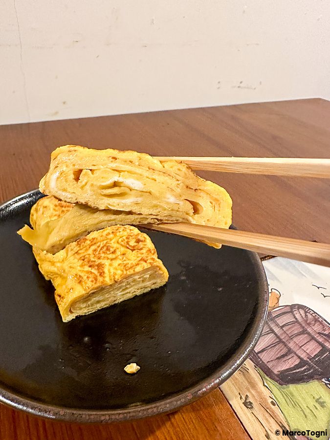 Un tamagoyaki sollevato con bacchette di legno sopra un piatto nero al Ristorante Momose Shokudo.