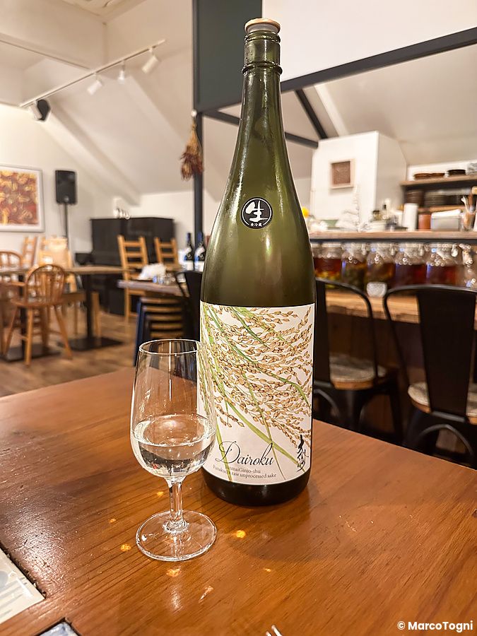 Una bottiglia di sakè Dairoku e un calice di vetro su un tavolo di legno al Ristorante Momose Shokudo.