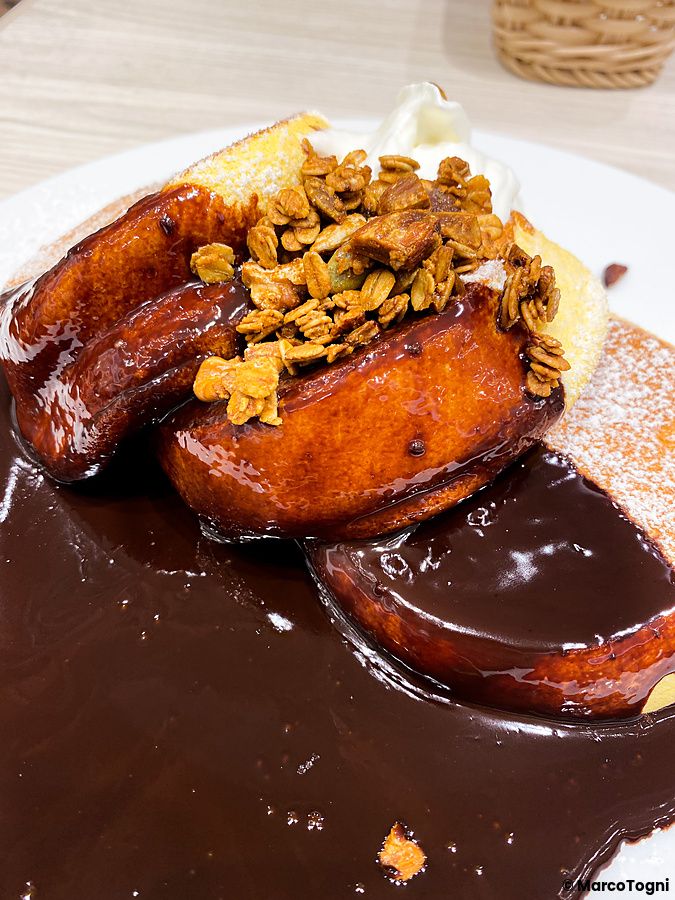 Un a Happy Pancake servito su un piatto bianco, ricoperto da una ricca salsa al cioccolato, granola e panna montata.