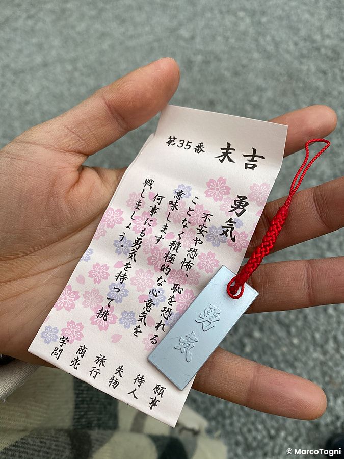 Una mano tiene un oracolo cartaceo con motivi floreali e un ciondolo metallico al santuario Kitaguchi Hongu Fuji Sengen.