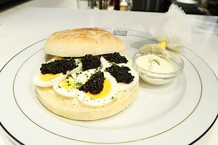 Un panino di lusso con caviale e uova sode su un piatto con logo Caviar House & Prunier accanto a una salsa bianca.