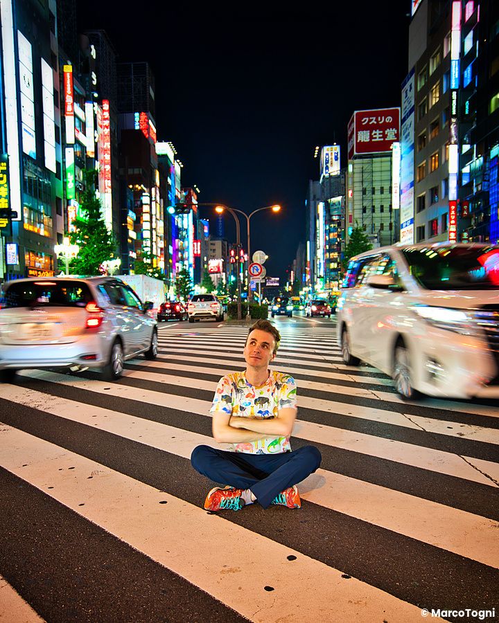 Marco Togni seduto al centro di un attraversamento pedonale illuminato dai neon nel quartiere di Shinjuku a Tokyo.