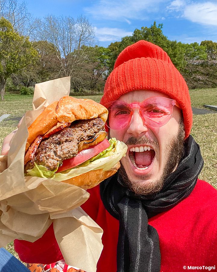 Marco Togni con cappello rosso tiene un burger al parco Tachikawa Kinen Koen.