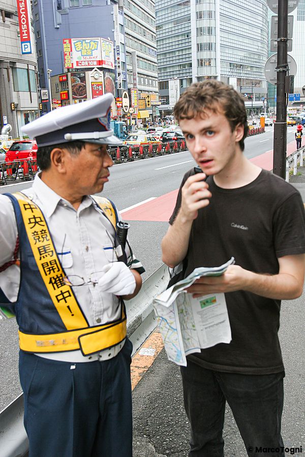 Un agente di polizia in uniforme parla con Marco Togni che consulta una mappa lungo una strada a Shinjuku.