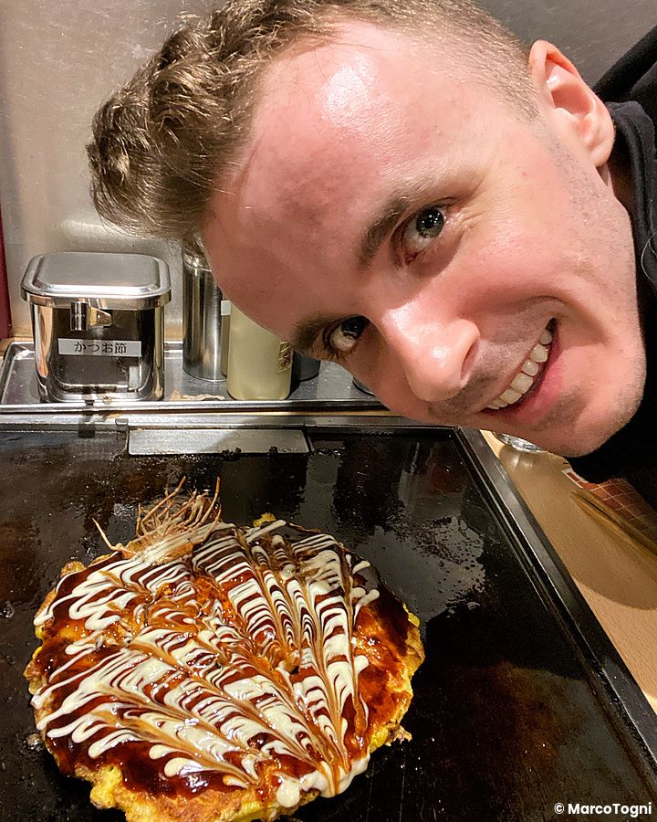 Marco Togni sorridente posa accanto a un okonomiyaki caldo su una piastra in un ristorante Chibo.