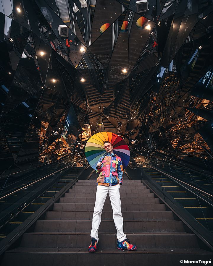 Marco Togni con ombrello arcobaleno in posa sulla scala mobile specchiata del Tokyu Plaza ad Harajuku.