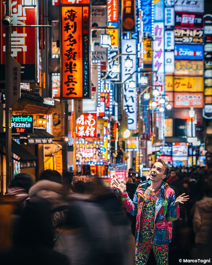 Marco Togni dai vestiti colorati guarda verso l'alto nel mezzo di una strada notturna illuminata dai neon a Shinjuku.