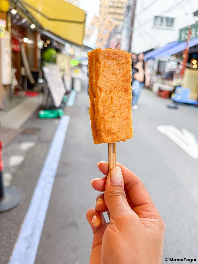 Una mano sostiene uno stecco con un tamagoyaki davanti alla via del mercato di Tsukiji, comprato da Yamacho.
