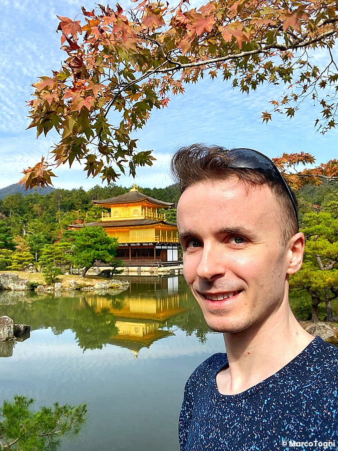 Marco Togni sorridente davanti al tempio d'oro di Kyoto con foglie d'autunno.