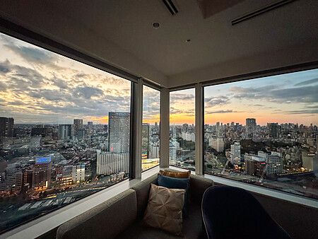 Una stanza d'hotel con ampie vetrate ad angolo che offrono una vista panoramica sullo skyline di Tokyo al tramonto.