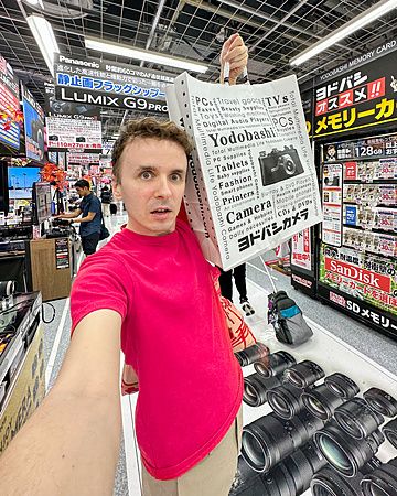 Marco Togni con busta di Yodobashi Camera in un negozio di elettronica.