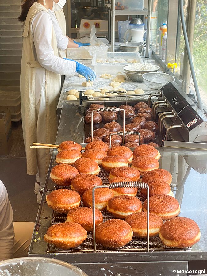Una persona prepara l'impasto per i donut di I'm donut ? in un laboratorio a vista con ciambelle fritte in primo piano.