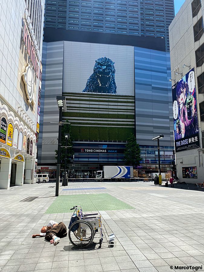 Una persona è distesa a terra accanto a una sedia a rotelle in una piazza a Kabukicho con l'edificio Toho Cinemas sullo sf...