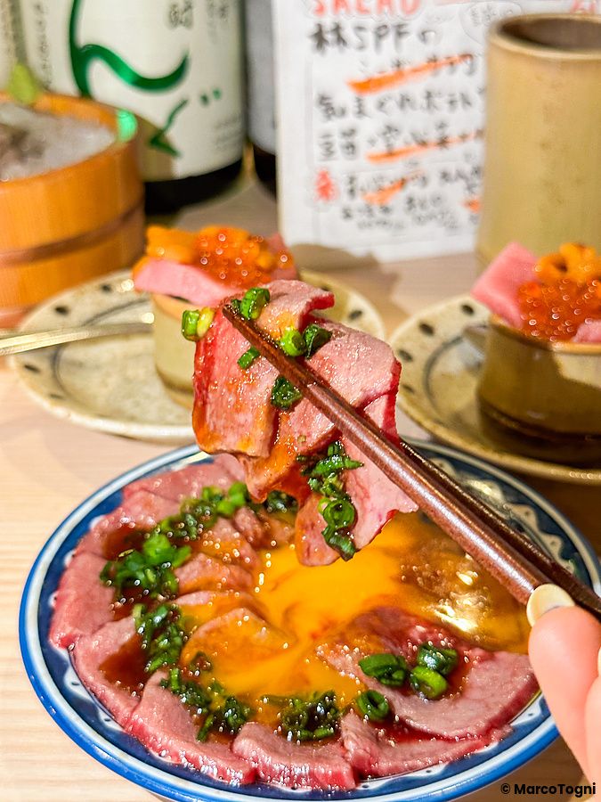 Una mano usa le bacchette per sollevare una fetta di manzo crudo a Shibuya Kito Kito in un piatto con tuorlo d'uovo.