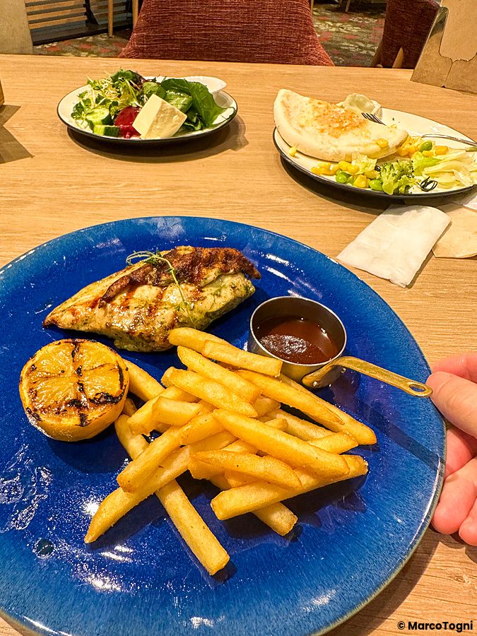 Un piatto blu con pollo e patatine a Sizzler accompagnato da una ciotolina di salsa su un tavolo in legno.