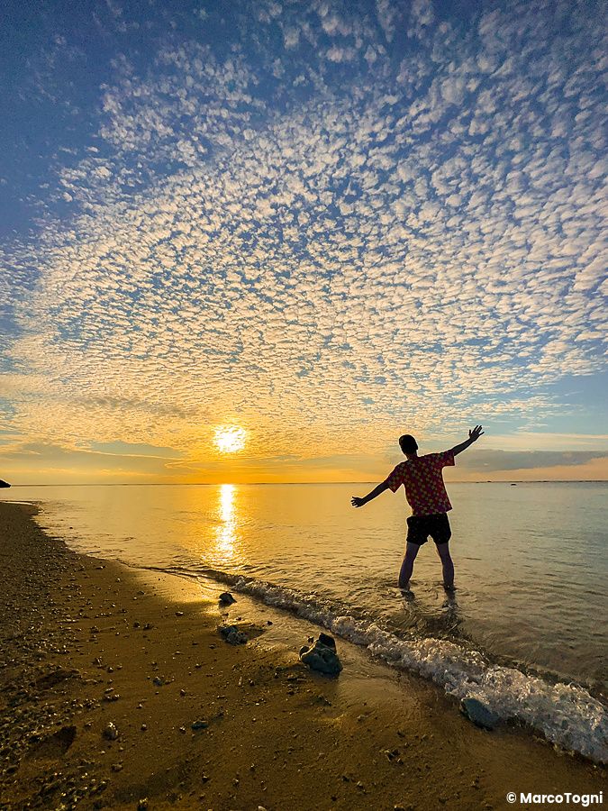 Una persona con le braccia aperte nell'acqua della spiaggia Mimikiri, a Iriomote, durante un tramonto dorato.