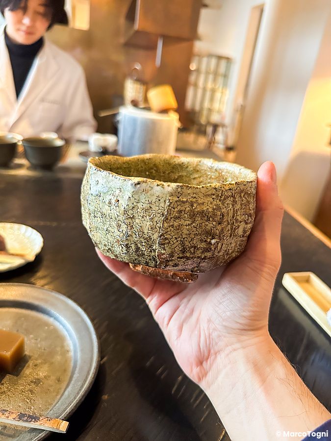 Una mano tiene una ciotola di ceramica ruvida e crepata al Sakurai Japanese Tea Experience, con un dolce sullo sfondo.