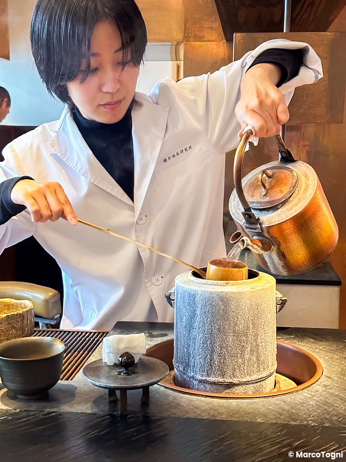 Una donna in camice bianco versa acqua calda da un bollitore in rame durante una preparazione al Sakurai Japanese Tea Expe...