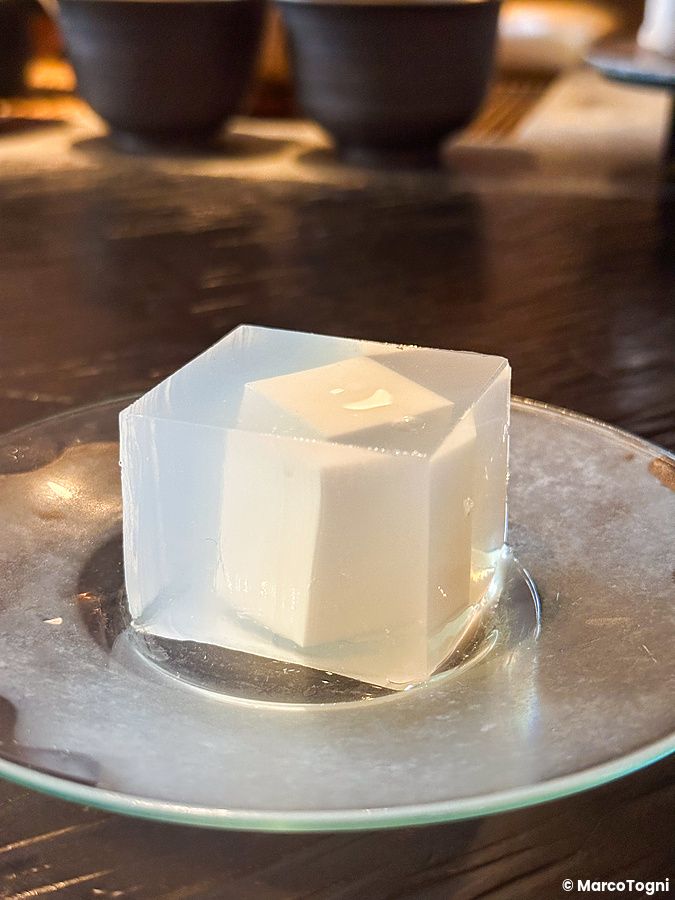 Un wagashi cubico traslucido con un centro solido su un piattino di vetro al Sakurai Japanese Tea Experience.